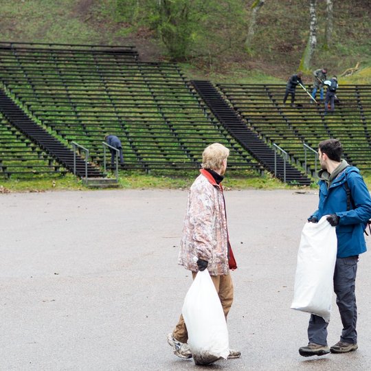 Du vyrai stovi lauko estrados aikštelėje, kiekvienas rankoje laiko baltą šiukšlių maišą. Jie atsisukę į šlaitą su mediniais suolais, kur matyti daugiau talkininkų, kurie grėbia lapus ir tvarko teritoriją. Vienas iš vyrų vilki mėlyną striukę, kitas – margą
