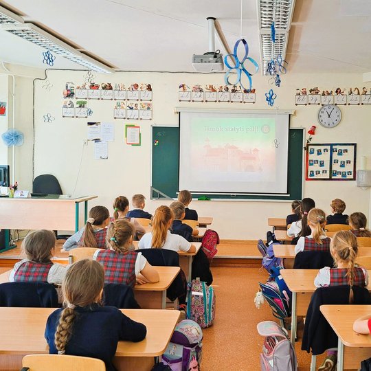 Pradinukai klasėje sėdi prie stalų ir žiūri į ekraną, kuriame rodomas skaidrių pristatymas su pavadinimu „Išmok statyti pilį!“. Klasė dekoruota raidžių plakatais ir kabančiomis mėlynomis dekoracijomis.