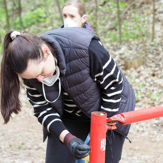 Moteris su pirštinėmis valo raudonai baltą metalinį užtvarą miško take, šalia jos kitas asmuo stebi procesą. Abi dėvi apsaugines kaukes, viena jų nuleidusi po smakru.