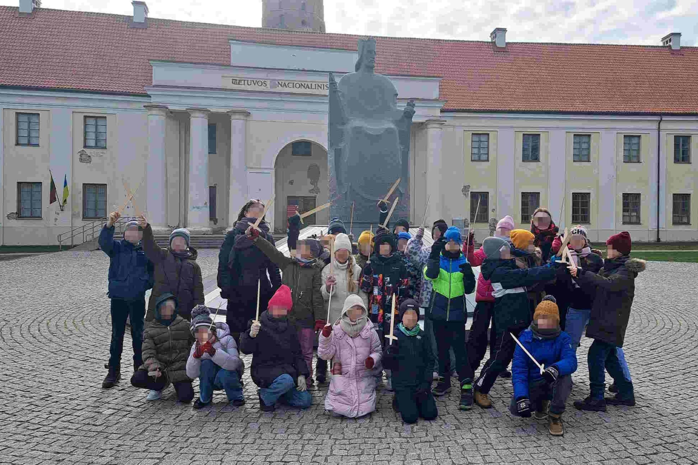 Nuotraukoje matome grupę vaikų Vilniaus pilių teritorijoje, priešais Lietuvos Nacionalinį muziejų. Jie sužavėti šios vietos istorija ir simboliu – Mindaugo paminklu. Vaikai turi suvenyrinius kalavijus, kurie suteikia jiems galimybę aktyviau įsijausti į is