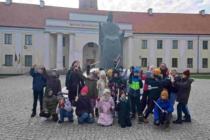 Nuotraukoje matome grupę vaikų Vilniaus pilių teritorijoje, priešais Lietuvos Nacionalinį muziejų. Jie sužavėti šios vietos istorija ir simboliu – Mindaugo paminklu. Vaikai turi suvenyrinius kalavijus, kurie suteikia jiems galimybę aktyviau įsijausti į is