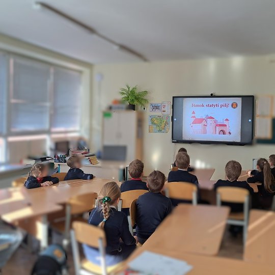 Maža vaikų grupė klasėje stebi skaidrę su užrašu „Išmok statyti pilį!“. Vaikai sėdi prie suolų, kambaryje daug šviesos, šone matyti žemėlapis ir mokytojos darbo vieta.