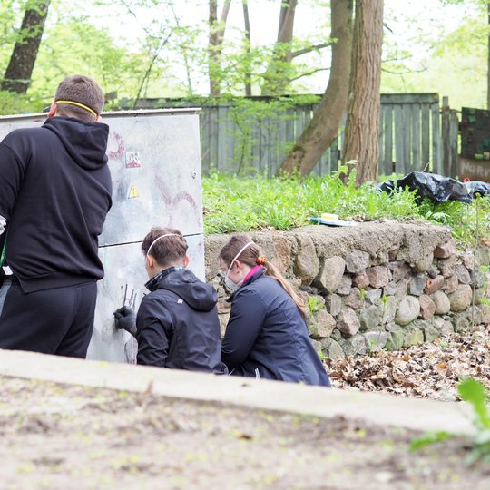 Trys žmonės valo grafičiais išteptą elektros skydinę, du jų pritūpę, o vienas stovi šalia. Aplinka žalia, matosi akmeninė atraminė siena ir miškinga teritorija.