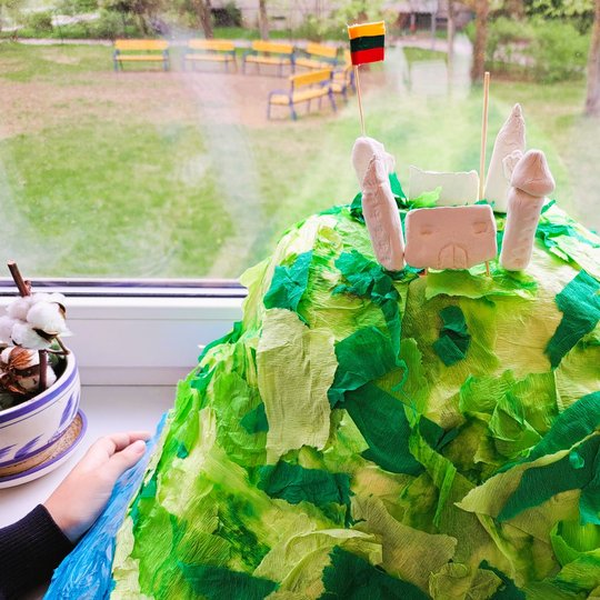 Vaikų sukurtas piliakalnio maketas padėtas ant palangės. Piliakalnis dengtas žaliomis ir gelsvomis krepinio popieriaus skiautėmis, viršuje pastatyta balta molinė pilis su bokštais ir vėliava – Lietuvos trispalve. Aplink langą matomas mokyklos kiemas su su