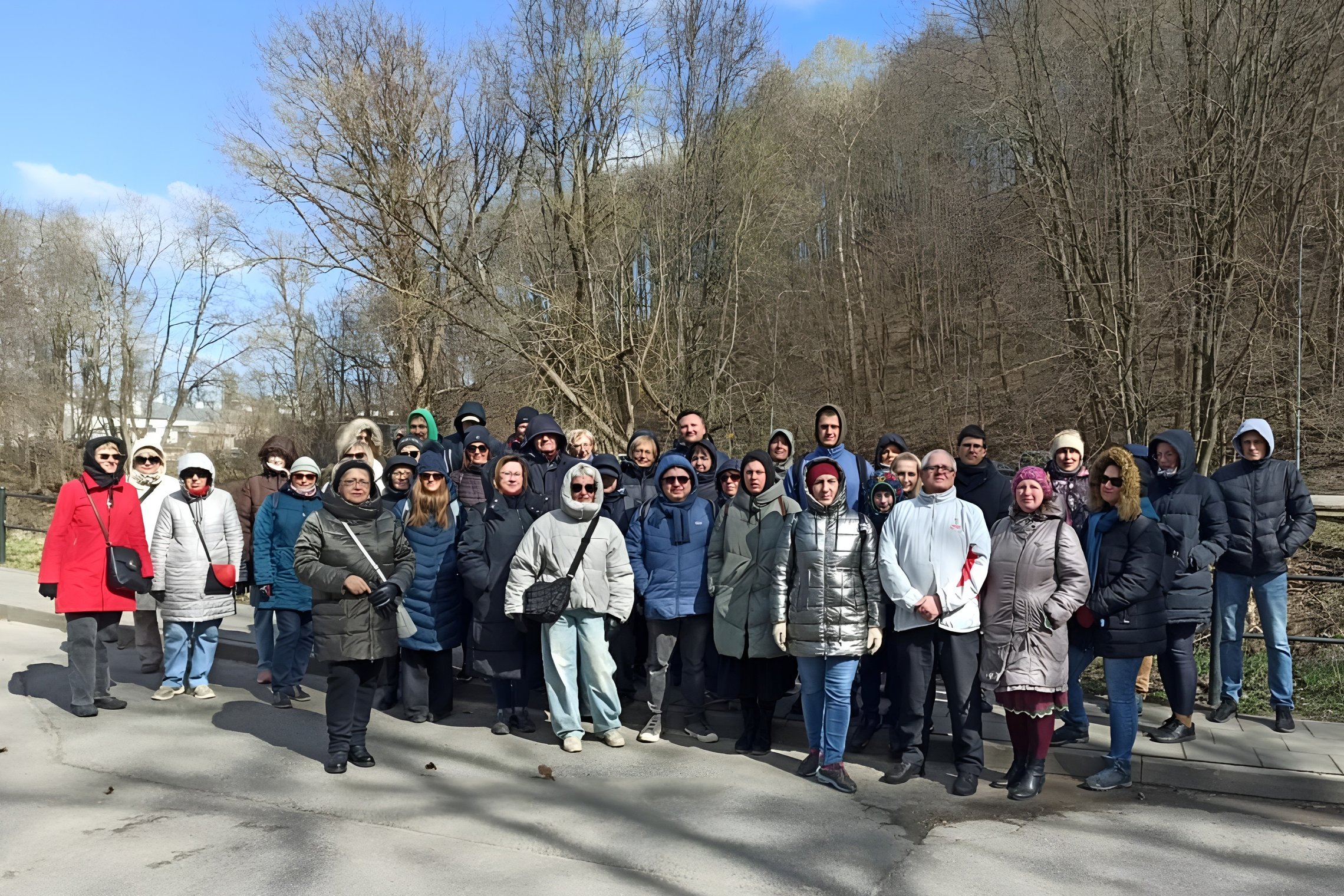 Grupė žmonių, dalyvavusių sekmadienio ekskursijoje, fotografuojasi kartu ant Vilnios kranto. Ekskursijos dalyviai susirinkę saulėtą, bet šaltą dieną, nes apsirengę šiltai. Už jų nugaros yra kalnas, piliakalnis, apaugęs medžiais.