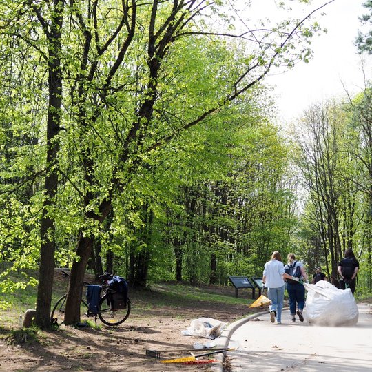 Grupė žmonių traukia didelį baltą maišą parko taku, šalia guli įrankiai ir dviratis atsirėmęs į medį. Aplink juos gausu žalios lapijos ir tankus pavasarinis miškas.