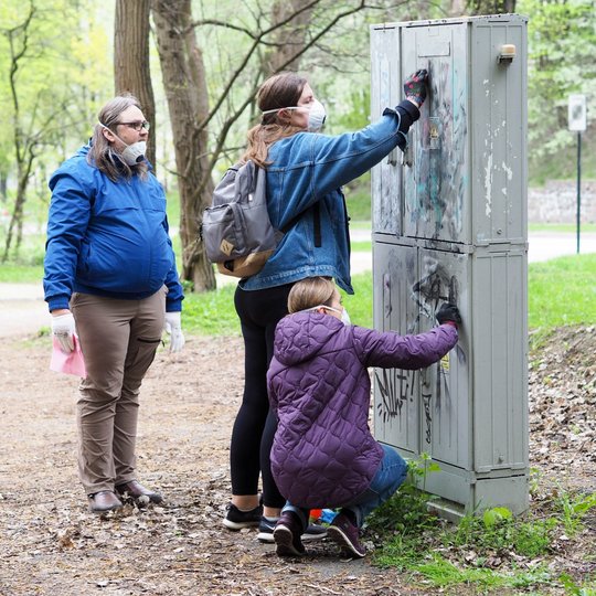 Dvi moterys valo grafičiais apipaišytą elektros skydinę parke, viena stovi, kita siekia viršų, o trečioji pritūpusi dirba prie apatinės dalies. Šalia jų stovi vyras su pirštinėmis, laikantis šluostę ir stebintis darbą.