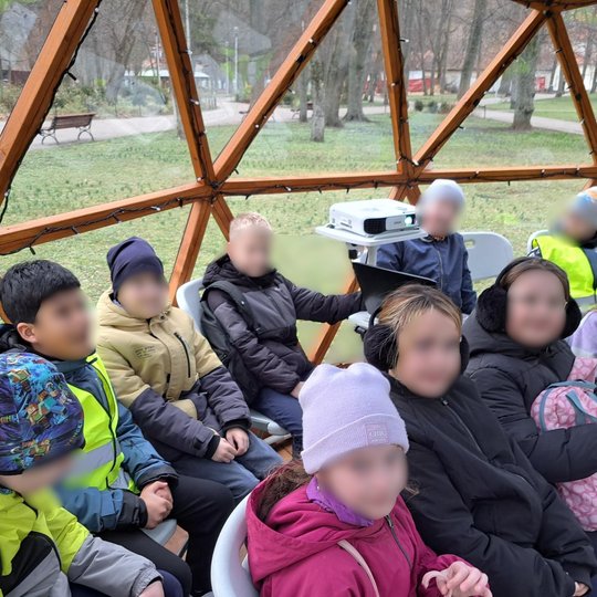Grupė vaikų sėdi geodezinio kupolo viduje ant baltų kėdžių, apsirengę šiltais drabužiais. Centre stovi projektorius ant stovo, ruošiamas vaizdo medžiagos rodymui. Kupolo langai atveria vaizdą į parką su takeliais, suolais ir medžiais. Vaikai atrodo susika
