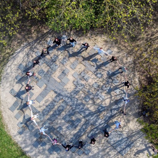Žmonės stovi ratu susikibę rankomis, vaizdas užfiksuotas iš viršaus. Jie stovi ant aikštelės su geometriniu raštu, apsuptos medžių žalumos.
