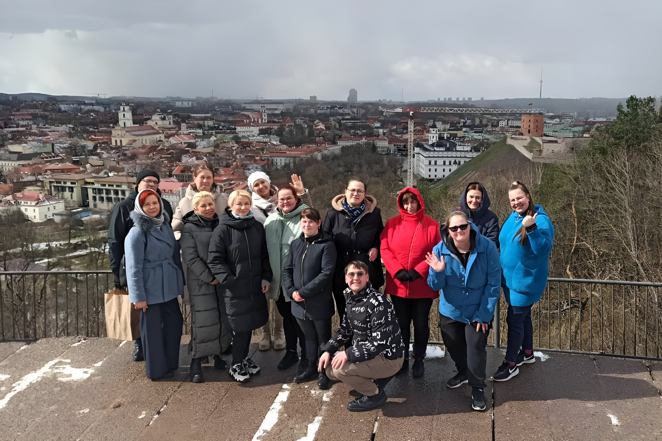 Grupė žmonių, susirinkusių ant aukšto Trijų Kryžių kalno, fone matosi Vilniaus miesto panorama ir Vilniaus pilys. Visi džiugiai mojuoja į kamerą. Ekskursijos dalyviai dėvi įvairius žiemos drabužius, nuo paltų iki striukių. Nors saulėta, bet šaltoka, žmonė