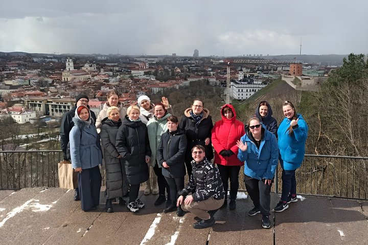 Grupė žmonių, susirinkusių ant aukšto Trijų Kryžių kalno, fone matosi Vilniaus miesto panorama ir Vilniaus pilys. Visi džiugiai mojuoja į kamerą. Ekskursijos dalyviai dėvi įvairius žiemos drabužius, nuo paltų iki striukių. Nors saulėta, bet šaltoka, žmonė