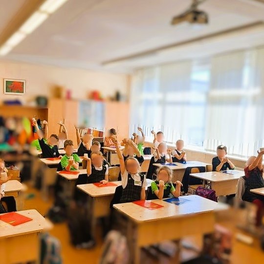 Pradinės mokyklos klasės nuotrauka, kurioje mokiniai sėdi prie savo suolų ir laiko rankose pakeltus molinius dirbinius. Vaikai vilki mokyklines uniformas, klasėje šviesu, pro langus sklinda dienos šviesa. Ant suolų padėtos spalvotos lentelės darbui, o fon