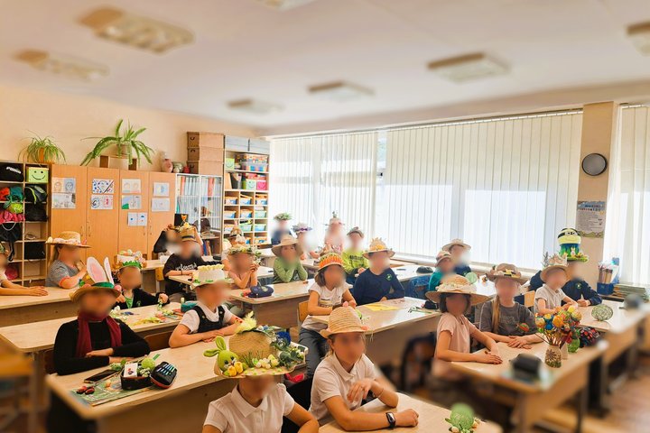 Nuotraukoje matoma mokyklos klasė, kurioje sėdi vaikai. Visi mokiniai dėvi kūrybiškai papuoštas skrybėles – ant jų galima pamatyti įvairius pavasario ar Velykų simbolius, tokius kaip kiškučiai, kiaušiniai, gėlės, žolė. Klasės aplinka spalvinga ir jauki: a