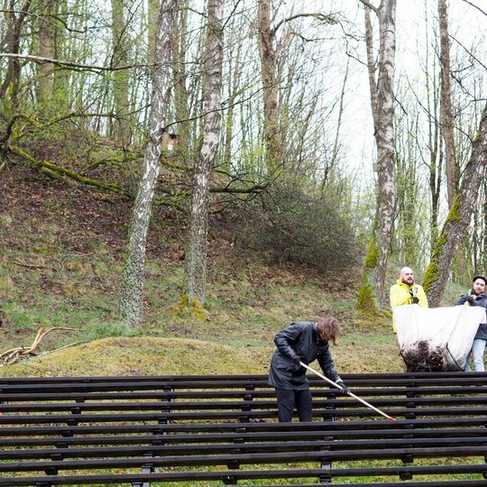 Lauko estradoje tarp beržų du žmonės valo suolus, o kiti du laiko didelį šiukšlių maišą su lapais. Aplink matyti pavasarėjantis miškas ir žaliuojantis šlaitas.