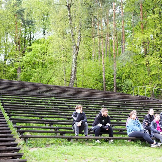 Grupė žmonių sėdi ant medinių lauko tribūnų miško apsuptyje, o vienas vyras su kuprine stovi priešais juos ir kalba. Aplinka žalia ir tankiai apaugusi medžiais, jaučiama pavasario atmosfera.