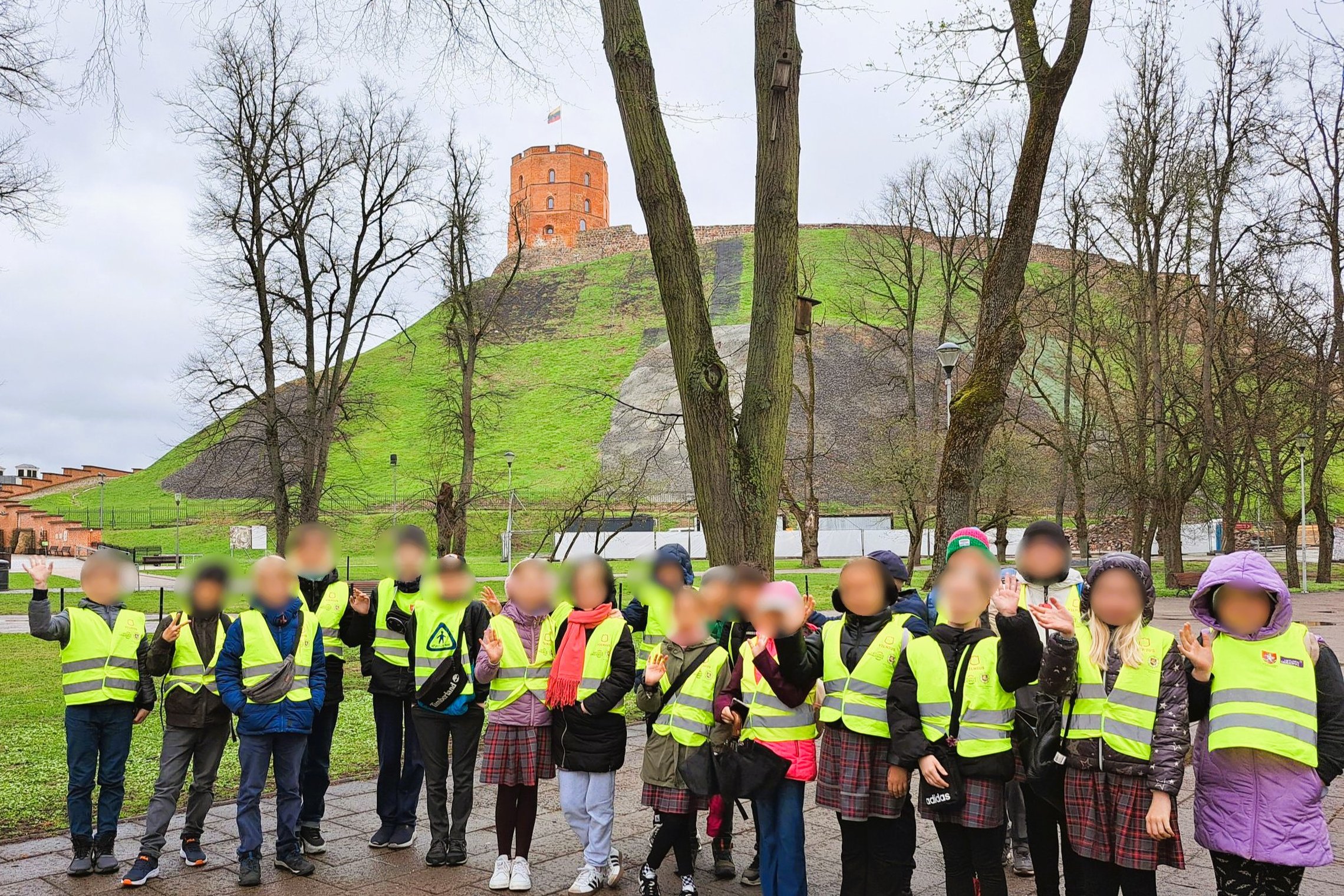 Vaikų grupė su ryškiaspalvėmis atšvaitinėmis liemenėmis stovi priešais Gedimino pilies bokštą Vilniuje, žaliuojančio kalno papėdėje, apsiniaukusią dieną. Kai kurie vaikai mojuoja.