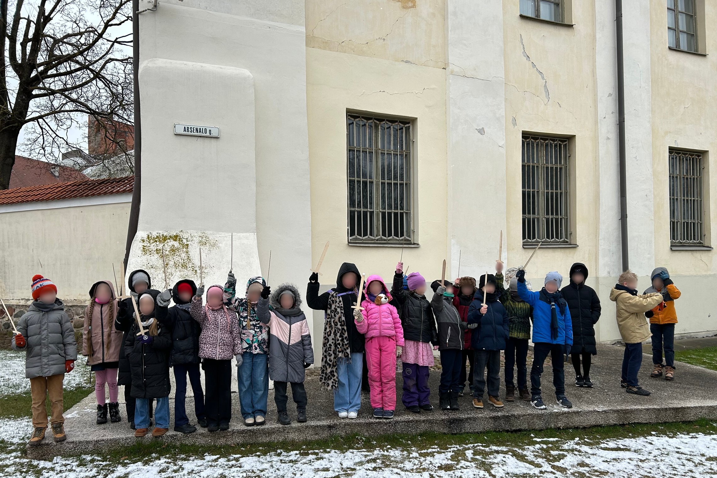 Grupė pradinukų stovi prie pastato, pažymėto gatvės ženklu „Arsenalo g.“, dalyvauja edukacinėje veikloje lauke. Vaikai šiltai apsirengę, rankose laiko medinius kalavijus. Už jų – Lietuvos nacionalinio muziejaus pastatas su grotomis dengtais langais. Ant ž