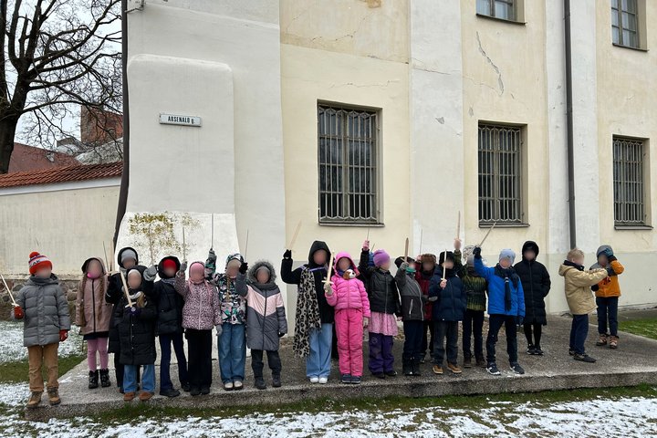 Grupė pradinukų stovi prie pastato, pažymėto gatvės ženklu „Arsenalo g.“, dalyvauja edukacinėje veikloje lauke. Vaikai šiltai apsirengę, rankose laiko medinius kalavijus. Už jų – Lietuvos nacionalinio muziejaus pastatas su grotomis dengtais langais. Ant ž