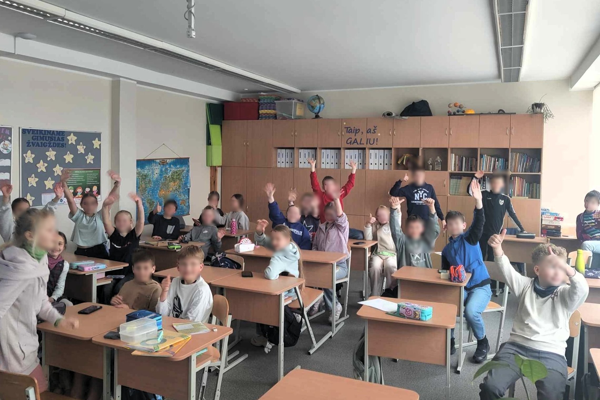 Mokyklos klasėje grupė vaikų entuziastingai kelia rankas, norėdami atsakyti į klausimą. Vaikai įvairiai apsirengę, sėdi prie medinių suolų šviesiame kambaryje su dideliais langais, pro kuriuos matyti žali medžiai.