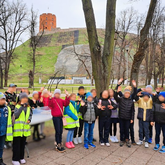 Grupė vaikų stovi prie lauko stalų su dėžėmis, iškėlę rankas ir šypsosi prie Gedimino pilies kalno Vilniuje. Dalis vaikų vilki ryškias geltonas saugos liemenes, kiti – šiltus rudens ar pavasario drabužius. Fone matomas Gedimino bokštas ir žalios šlaito te