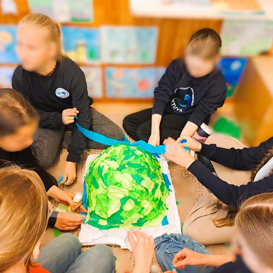 Grupė vaikų sėdi ratu ir kuria piliakalnio maketą iš įvairių žalių ir mėlynų popieriaus skiaučių. Centre – žalias kalnelis, atvaizduojantis piliakalnį. Vaikai įsitraukę į kūrybinį darbą, dirba komandoje. Fone – mokyklinės piešinių parodos ir plakatai ant 