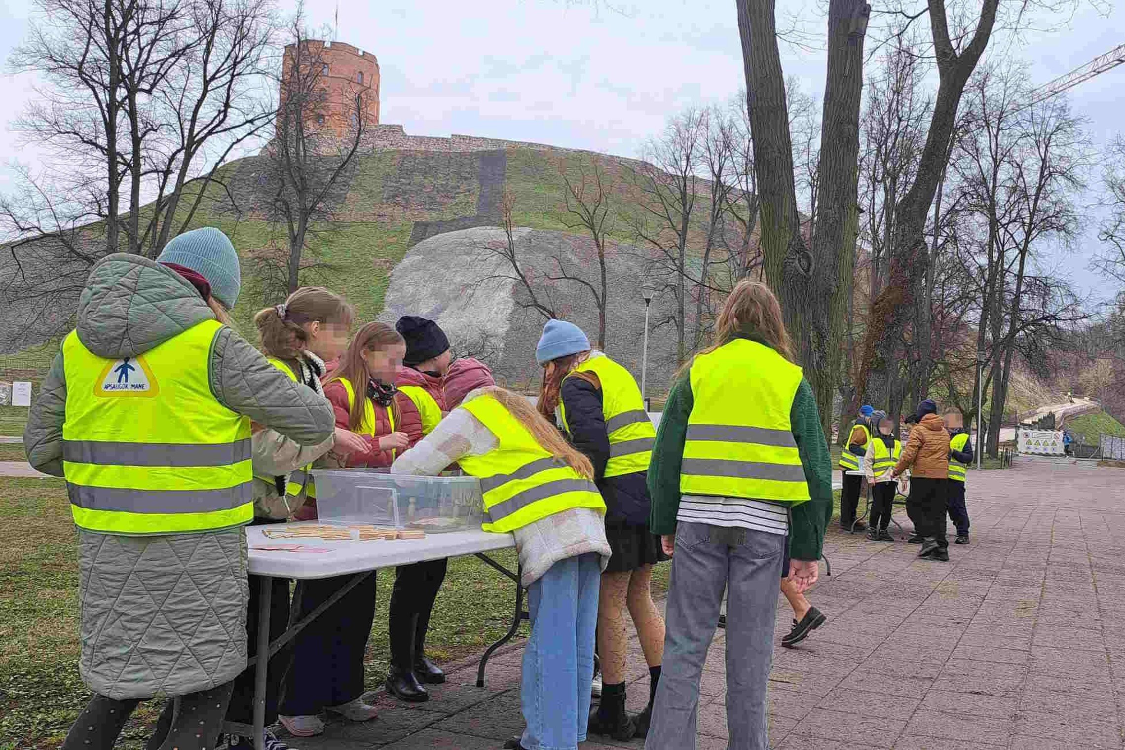 Grupė vaikų su ryškiai geltonomis saugos liemenėmis atlieka veiklą prie lauko stalų, pastatytų šalia Gedimino pilies kalno Vilniuje. Vaikai susikaupę tyrinėja ar konstruoja iš dėžėse esančių medžiagų. Fone matoma Gedimino pilies bokštas ir šlaitas, o oras