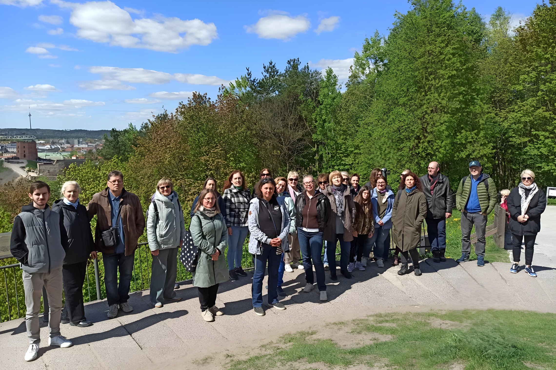 Ekskursijos „Vilniaus ritualai: plikieji kalnai, geležiniai šarvai“ dalyviai pozuoja nuotraukai su Vilniaus pilių panorama fone – matomas Gedimino pilies bokštas, pilys ir žalios kalvos.