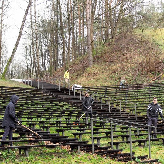 Lauko estrados suoliukuose, tarp medžių apsuptyje, keli žmonės atlieka talkos darbus – grėbia pernykščius lapus ir tvarko teritoriją. Kai kurie dėvi lietpalčius ar sportinius drabužius, o vienas asmuo tolumoje lipa laiptais tarp kalvų.