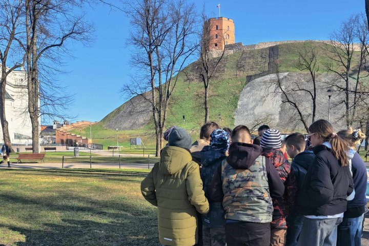 Grupė moksleivių stovi prie Gedimino kalno papėdės Vilniuje, atsukę nugaras į kamerą, žvelgdami į viršuje stūksančią Gedimino pilies bokšto raudonų plytų siluetą. Aplink – pavasariškas kraštovaizdis su plikais medžiais ir žaliuojančia kalno šlaite.