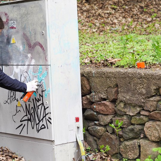 Moteris su pirštinėmis ir apsaugine kauke purškia valiklį ant grafičiais išmargintos elektros skydinės. Šalia matyti akmeninė atraminė siena ir nukritę lapai.