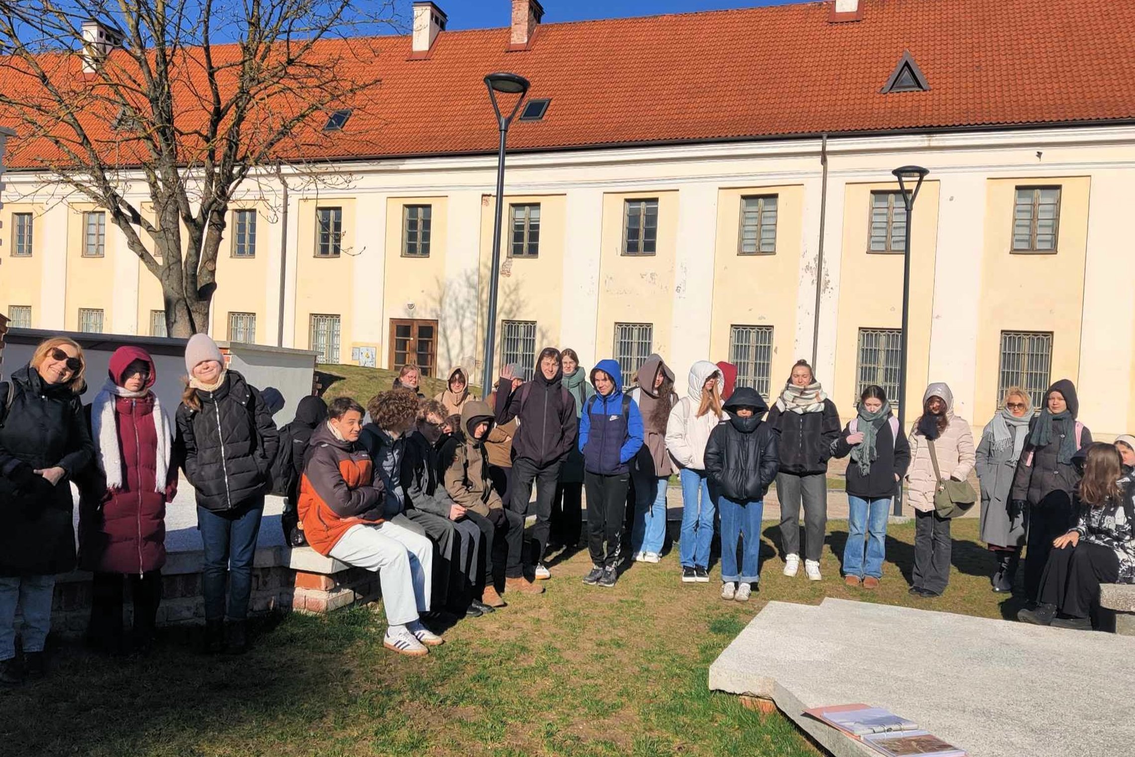 Devintokų grupė su mokytojais stovi ir sėdi prie aikštelės Vilniaus pilių teritorijoje, saulėtą, bet šaltą rytą. Fone – istorinis pastatas su raudonu stogu ir mažais langais. Visi šiltai apsirengę, su kepurėmis, šalikais ir striukėmis, kai kurie šypsosi. 