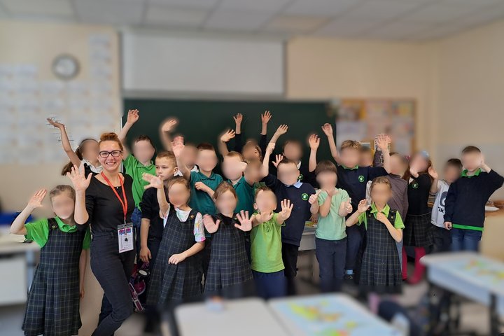 Grupinė nuotrauka klasėje, kurioje stovi besišypsanti edukatorė su mokiniais. Vaikai dėvi mokyklines uniformas – žalias arba tamsiai mėlynas palaidines, kai kurie – languotus sarafanus. Visi linksmai mojuoja rankomis, fone matyti lenta, laikrodis ir klasė