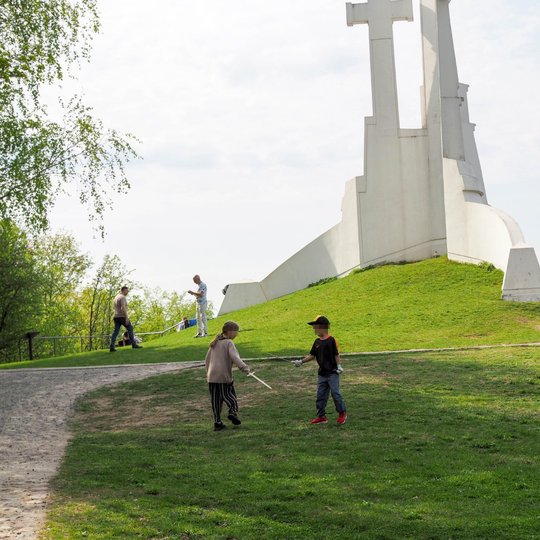 Vaikai žaidžia su lazdomis prie Trijų Kryžių paminklo ant žalios kalvos Vilniuje. Fone matyti daugiau žmonių, vaikštinėjančių ar užsiimančių veikla po atviru dangumi.