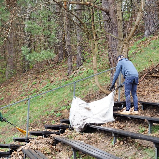 Du vyrai dirba lauko estradoje tarp medinių suolų, apsuptų miškingo šlaito. Vienas vyras stovi kairėje pusėje, vilkėdamas žalią striukę ir raudonus šortus, ir šluoja pernykščius lapus nuo laiptų.