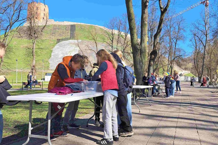 Nuotraukoje – vaikai dalyvauja lauko edukacijoje prie Gedimino kalno Vilniuje. Jie stovi prie kelių baltų stalų, sustatytų palei taką, ir aktyviai kažką veikia – galbūt tyrinėja, dėlioja ar kuria. Vaikai apsirengę lengvais pavasariškais rūbais, kai kurie