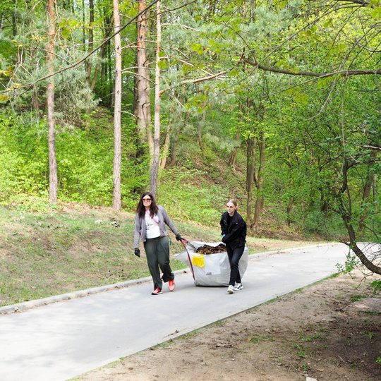 Dvi moterys traukia pilną lapų maišą asfaltuotu keliuku per pavasarėjantį mišką. Aplinkui žaliuoja medžiai, o šalia kelio matyti šlaitas ir tanki augmenija.