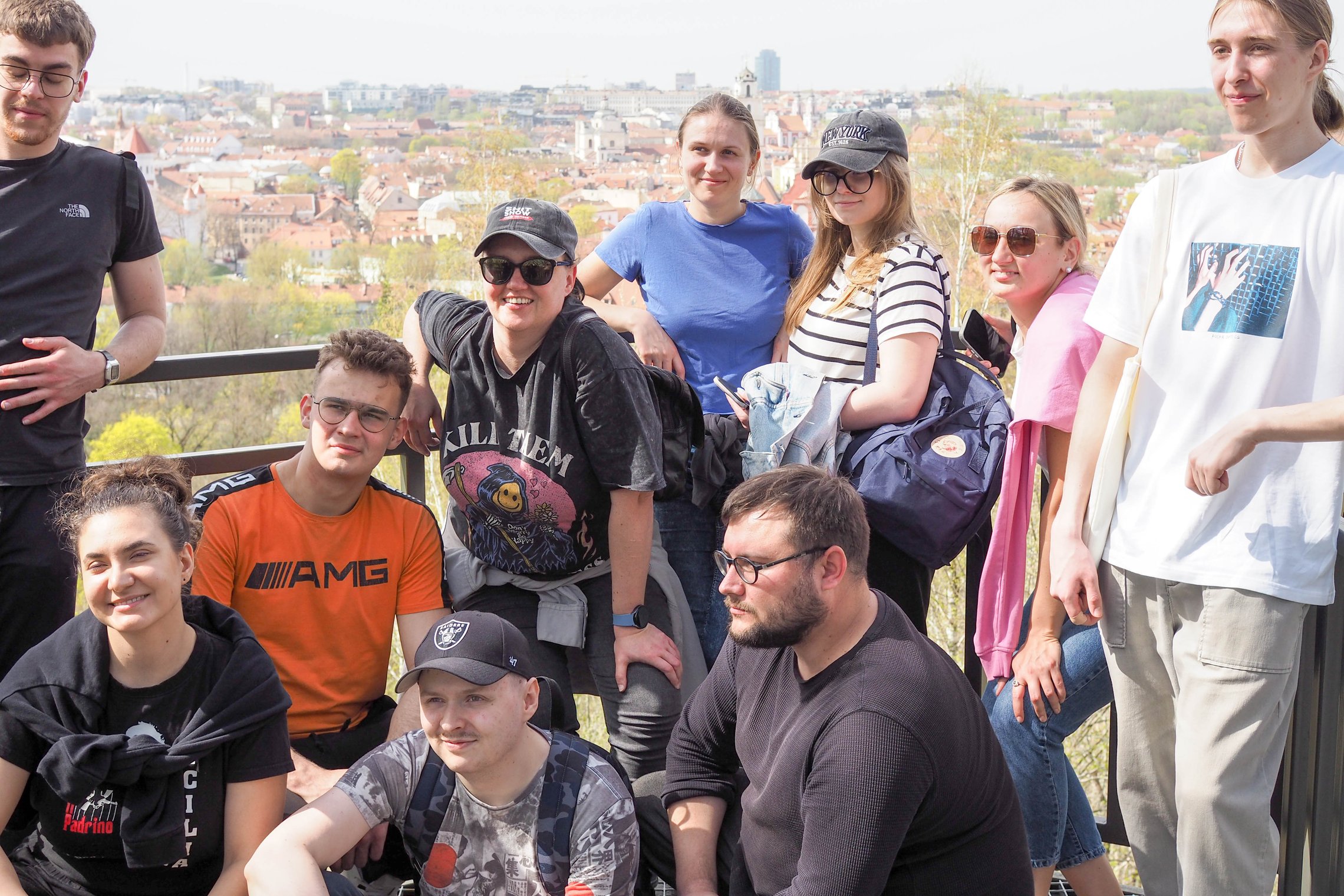 Grupė jaunų žmonių pozuoja nuotraukai ant apžvalgos aikštelės su vaizdu. Fone matyti raudoni senamiesčio stogai, bažnyčių bokštai ir modernūs pastatai tolumoje. Visi dalyviai šypsosi arba atsipalaidavę pozuoja, vilki laisvalaikio drabužius, kai kurie dėvi
