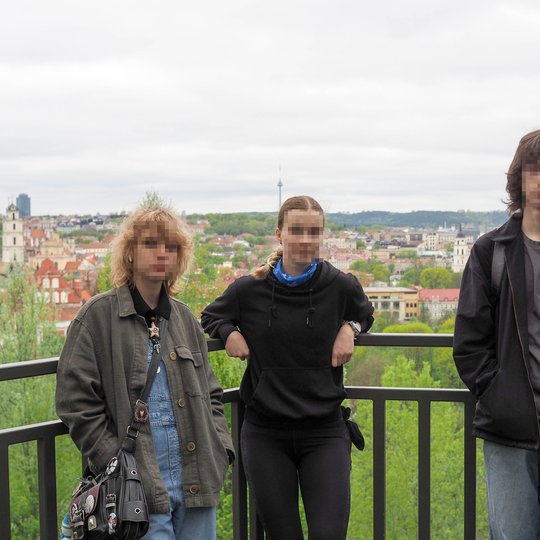 Trys jaunuoliai pozuoja ant apžvalgos aikštelės turėklų, už jų atsiveria Vilniaus senamiesčio panorama. Foną puošia raudoni stogai, bažnyčių bokštai ir tolumoje stovinti televizijos bokšto konstrukcija.