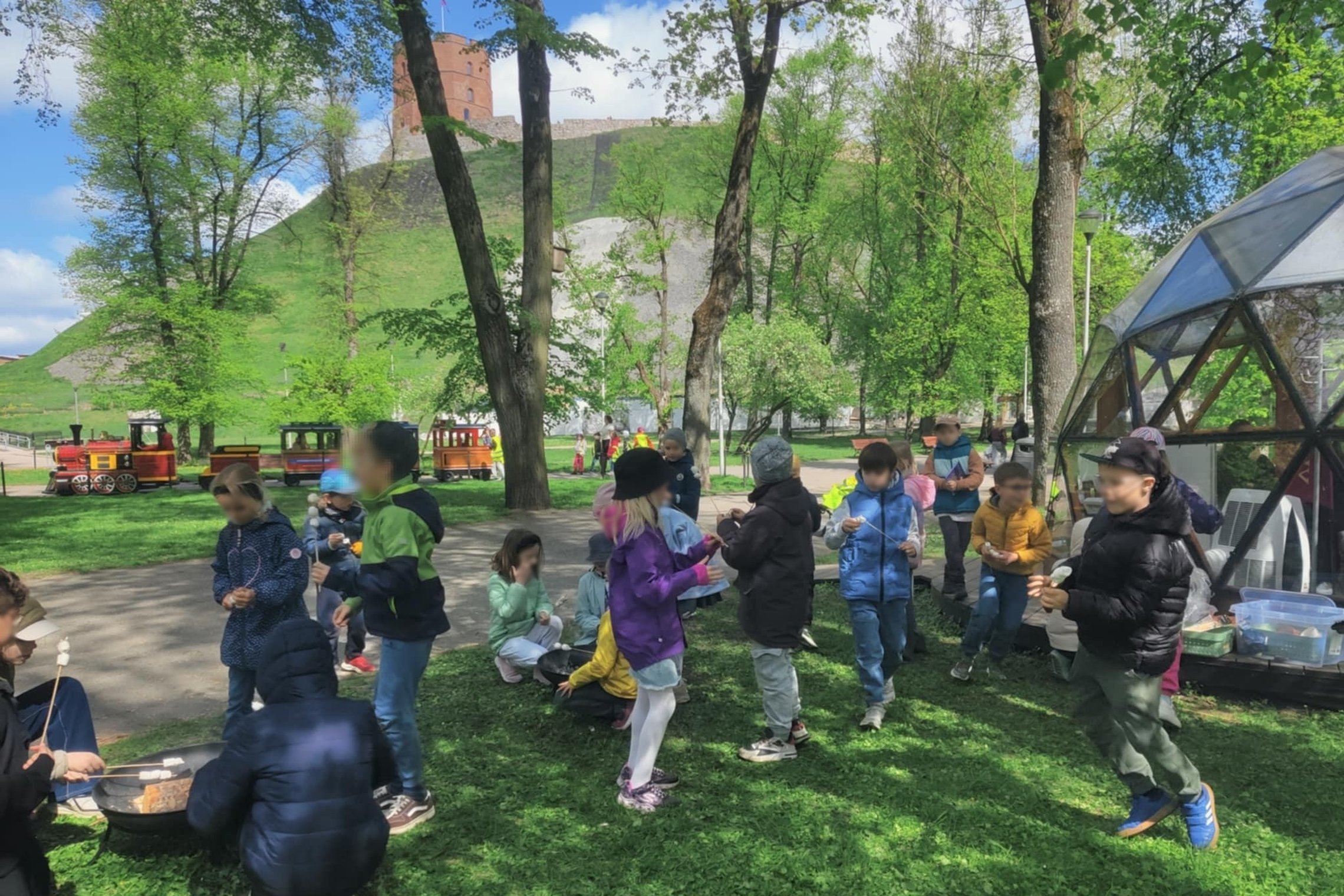 Vaikai kepa zefyrus lauke, šalia kupolo, medžių apsuptyje prie Gedimino kalno, kur matyti raudonų plytų Gedimino pilies bokšto viršūnė. Fone važiuoja spalvingas vaikiškas traukinukas.