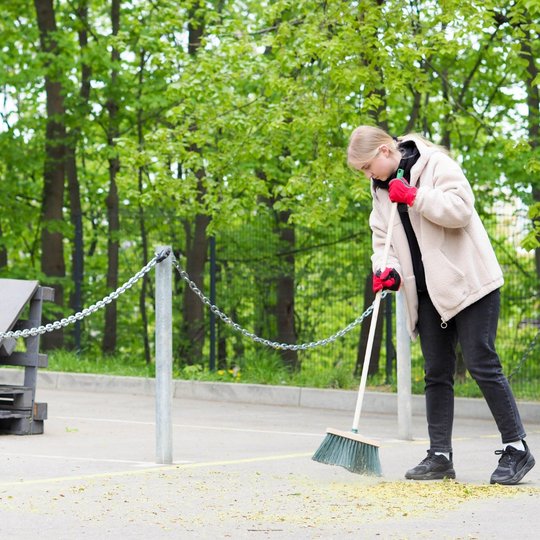 Moteris šluoja žiedadulkes ir smulkius lapus nuo asfaltuotos aikštelės, vilkėdama šiltą šviesų paltą ir ryškias raudonas pirštines. Aikštelė aptverta grandinėle, o fone stovi medžiai ir padėklai.