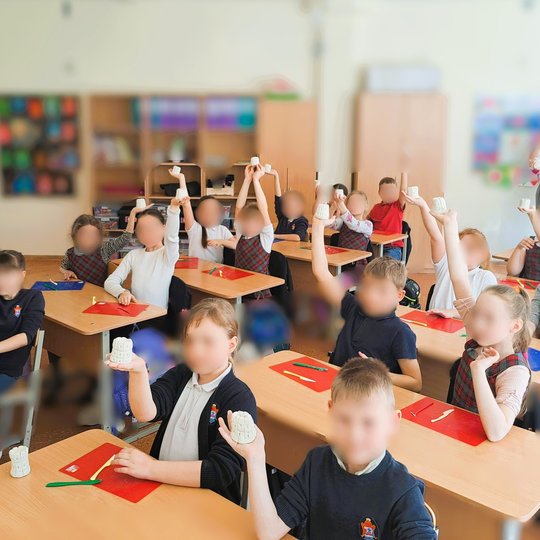 Pradinukai klasėje džiugiai kelia į viršų savo nulipdytas baltas molines pilis. Ant stalų padėti spalvoti padėkliukai ir molio formavimo įrankiai. Vaikai vilki uniformas languotais raštais.