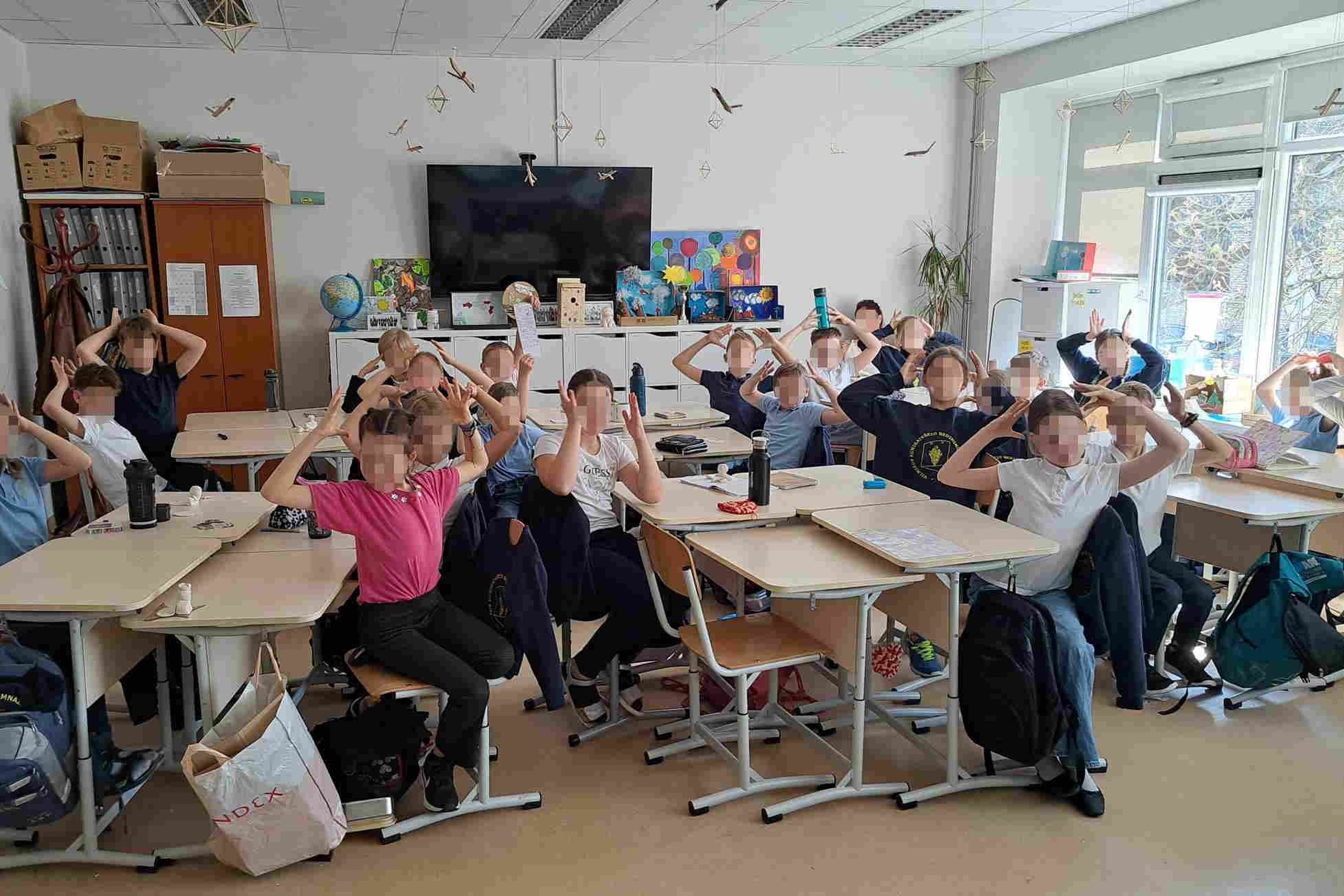 Trečios klasės mokiniai sėdi klasėje prie stalų ir dalyvauja edukacinėje veikloje, susijusioje su laisvės ir demokratijos tema. Vaikai pakėlę rankas virš galvos atlieka žaidybinę užduotį. Klasės aplinka dekoruota edukaciniais plakatais ir kūrybiniais darb