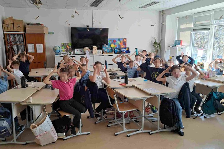 Trečios klasės mokiniai sėdi klasėje prie stalų ir dalyvauja edukacinėje veikloje, susijusioje su laisvės ir demokratijos tema. Vaikai pakėlę rankas virš galvos atlieka žaidybinę užduotį. Klasės aplinka dekoruota edukaciniais plakatais ir kūrybiniais darb