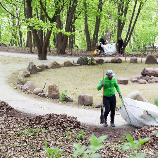 Miško aikštelėje su akmenų ratu žmonės valo aplinką – vienas asmuo priekyje tempia didelį baltą maišą su lapais. Tolumoje dar keli talkininkai krauna lapus į kitą maišą prie takelio.