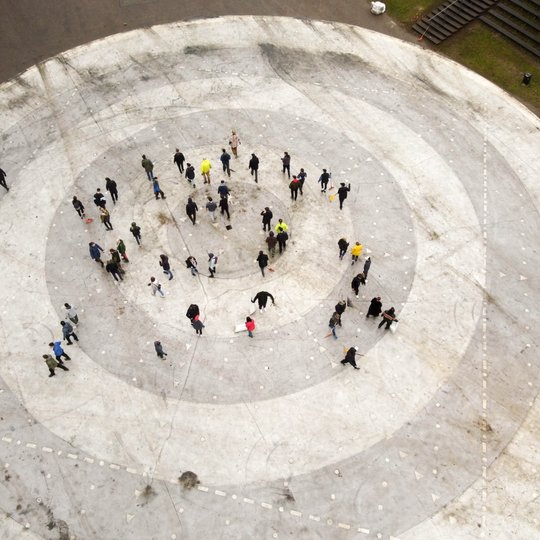 Nuotrauka iš viršaus rodo žmonių grupę, susibūrusią didelėje apskritimo formos aikštėje su koncentriškais raštais. Aikštė atrodo kaip lauko estrada ar viešoji erdvė renginiams. Žmonės išsisklaidę grupelėmis, kai kurie juda, o kiti stovi.