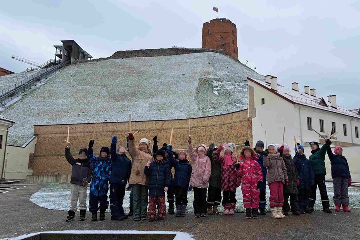 Grupė pirmokų stovi prie Gedimino pilies kalno Vilniuje, laikydami iškeltas medines kalavijų formos lazdeles. Fone – Gedimino pilies bokštas su plevėsuojančia Lietuvos vėliava. Žemę dengia lengvas sniego sluoksnis.