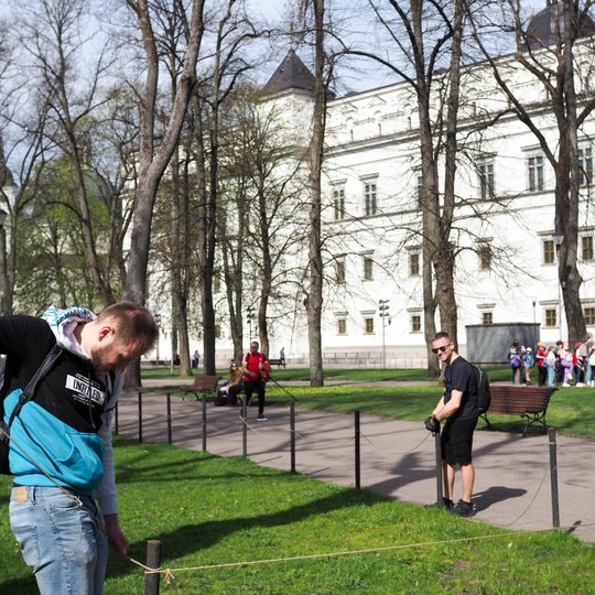 Du savanoriai tvarko virvinę tvorelę Sereikiškių parke, fone matyti Valdovų rūmai ir ekskursijoje dalyvaujanti vaikų grupė.