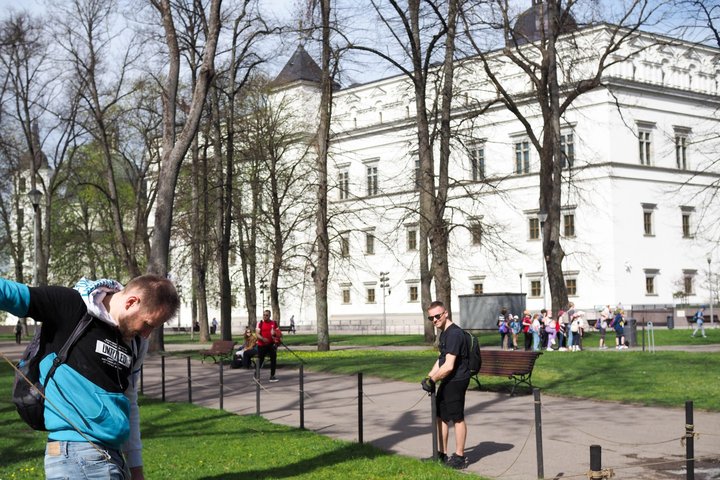 Du savanoriai tvarko virvinę tvorelę Sereikiškių parke, fone matyti Valdovų rūmai ir ekskursijoje dalyvaujanti vaikų grupė.