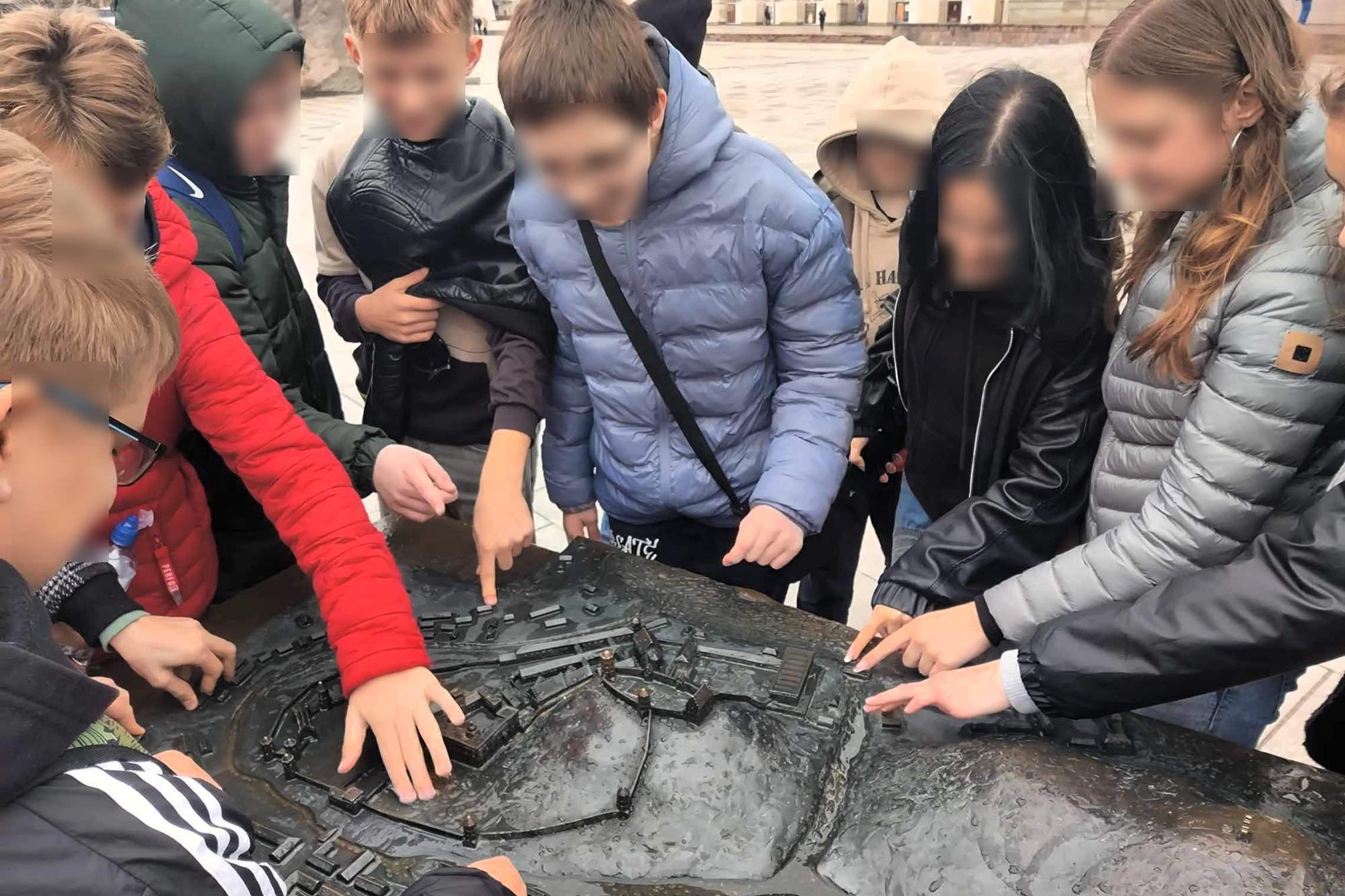 Grupė mokinių, apsirengusių šiltais pavasariniais drabužiais, susibūrė aplink bronzinį Vilniaus pilių maketą Katedros aikštėje. Vaikai rodo pirštais į skirtingas maketo vietas, tyrinėdami teritorijos struktūrą ir istorinius objektus. Fone matyti Katedros