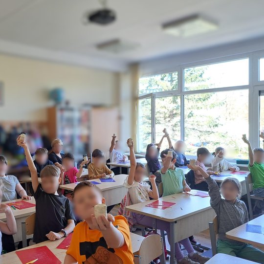 Pradinės mokyklos klasės nuotrauka, kurioje mokiniai sėdi prie savo suolų ir laiko rankose pakeltus molinius dirbinius. Vaikai vilki mokyklines uniformas, klasėje šviesu, pro langus sklinda dienos šviesa. Ant suolų padėtos spalvotos lentelės darbui, o fon